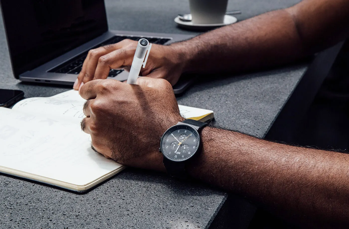 Mãos escrevendo em um caderno com caneta, relógio no pulso, notebook e xícara de café na mesa
