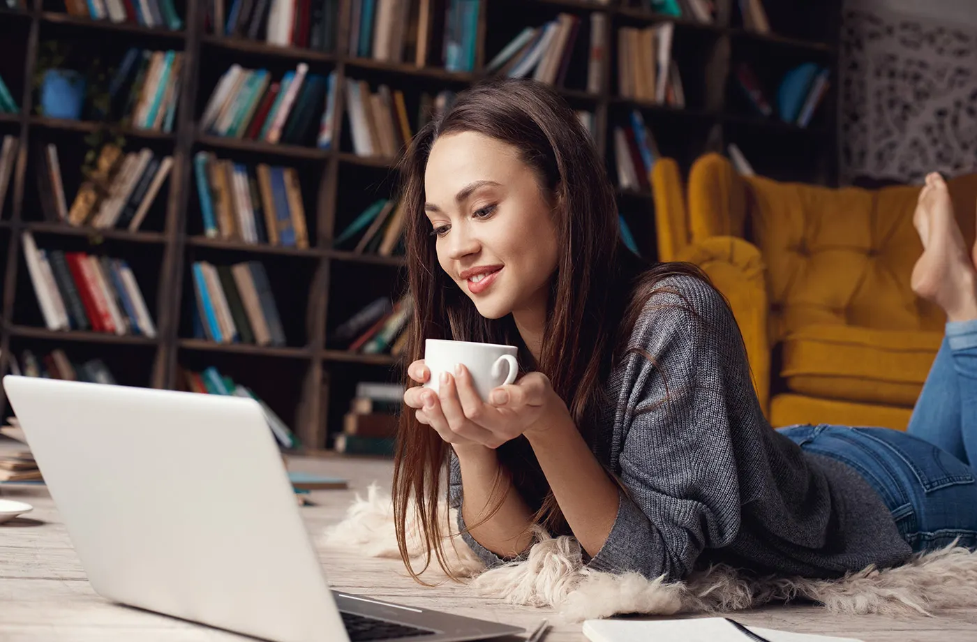Mulher deitada no chão com xícara de café em frente a um notebook, estante de livros ao fundo